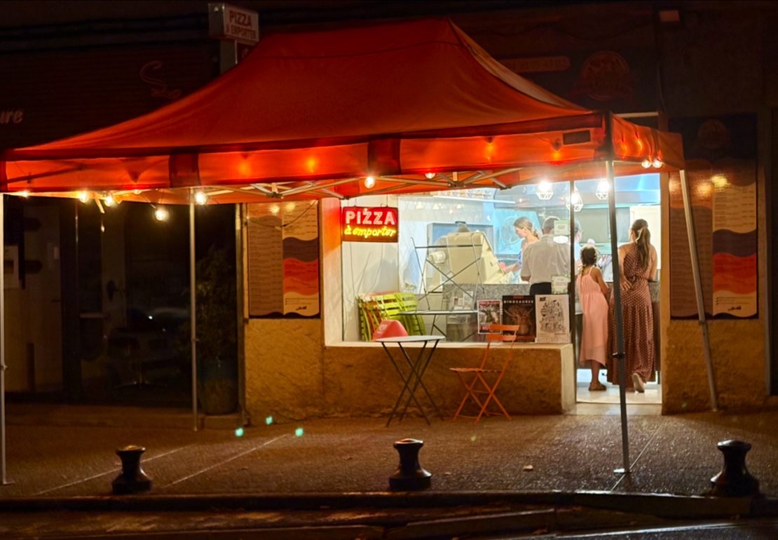 La devanture de la pizzeria de nuit : auvent rouge, guirlandes et enseigne « Pizza à emporter »